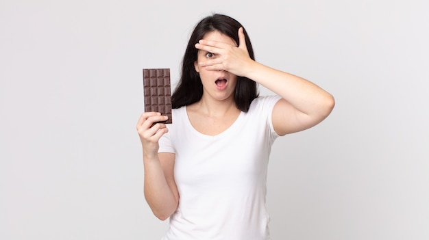 Mooie vrouw die geschokt, bang of doodsbang kijkt, haar gezicht bedekt met de hand en een chocoladereep vasthoudt