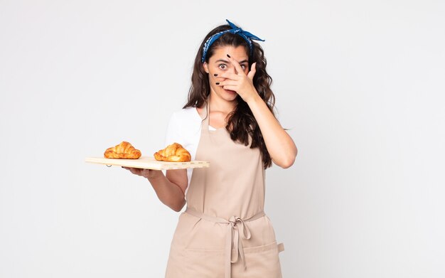 Mooie vrouw die geschokt, bang of doodsbang kijkt, haar gezicht bedekt met de hand en een dienblad met croissants vasthoudt