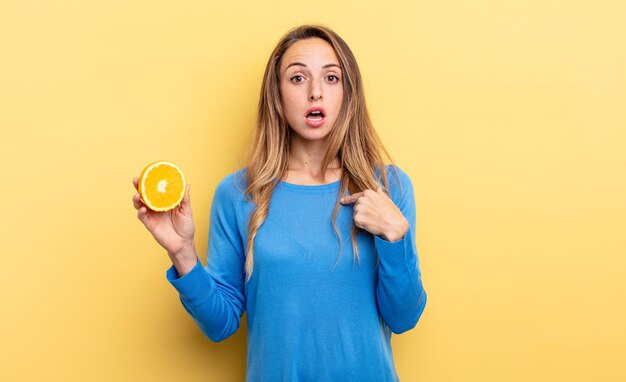 Mooie vrouw die geschokt en verrast kijkt met wijd open mond, wijzend naar zichzelf met half oranje