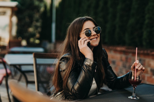 Mooie vrouw die in een café zit, een cocktail drinkt en een smartphone gebruikt. Student tijdens pauze in het café. Levensstijl, technologie