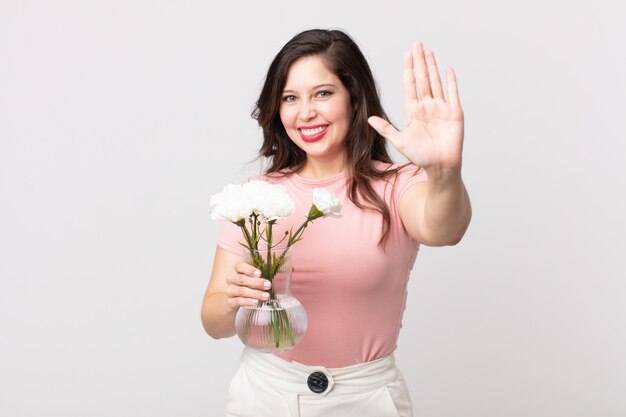 Mooie vrouw die lacht en er vriendelijk uitziet, nummer vijf toont en een decoratieve bloemenpot vasthoudt