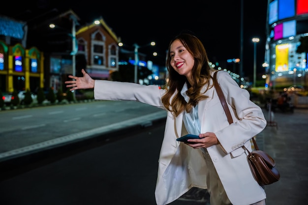 Mooie vrouw die lacht met behulp van smartphone applicatie begroeting met hand omhoog bellende taxi na laat werk, Aziatische zakenvrouw staande hagel zwaaiende handtaxi op weg in drukke stadsstraat 's nachts