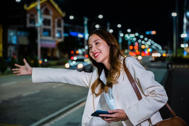 Mooie vrouw die lacht met behulp van smartphone applicatie begroeting met hand omhoog bellende taxi na laat werk, Aziatische zakenvrouw staande hagel zwaaiende handtaxi op weg in drukke stadsstraat 's nachts