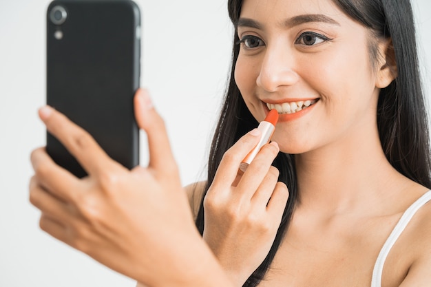 Mooie vrouw die lippenstift toepast terwijl ze haar mobiele telefoon als spiegel over een witte muur houdt. Aziatische vrouwelijke blogger