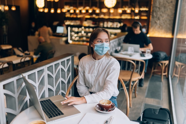 Mooie vrouw die medisch gezichtsmasker draagt dat laptop met behulp van om te werken