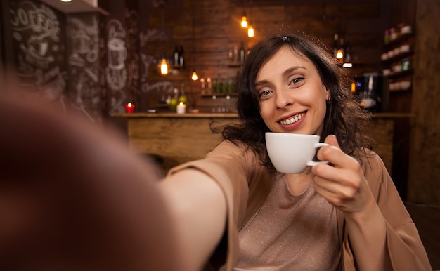 Mooie vrouw die mobiele telefoon gebruikt om een selfie te maken tijdens haar werk in een coffeeshop. Mooi meisje met een kopje koffie. Jonge vrouw ontspannen in een coffeeshop. Casual dag voor een jonge vrouw.