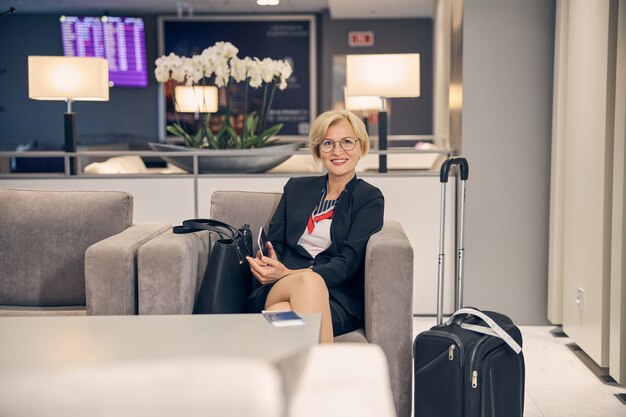 Mooie vrouw die mobiele telefoon vasthoudt en glimlacht terwijl ze in een fauteuil zit voor een zakenreis