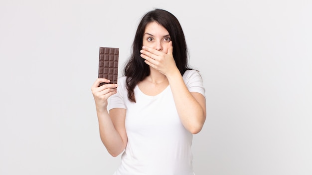 Mooie vrouw die mond bedekt met handen met een geschokte en een chocoladereep vasthoudt