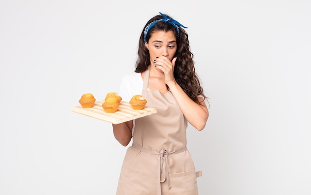 Mooie vrouw die mond bedekt met handen met een geschokte en een muffinsdienblad houdt