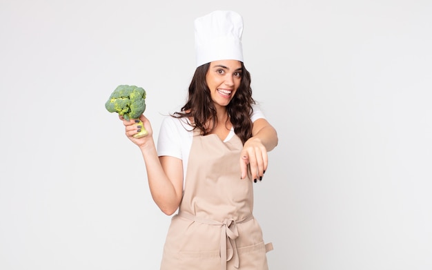 Mooie vrouw die naar de camera wijst en ervoor kiest dat je een schort draagt en een broccoli vasthoudt