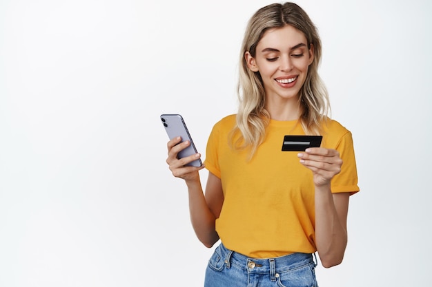 Mooie vrouw die online betaalt met creditcard en mobiele telefoon die op internet winkelt in gele t-shirt en jeans op witte achtergrond