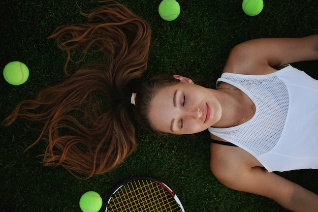 Mooie vrouw die op de grastennisbaan ligt met tennisballen om haar heen, bovenaanzicht