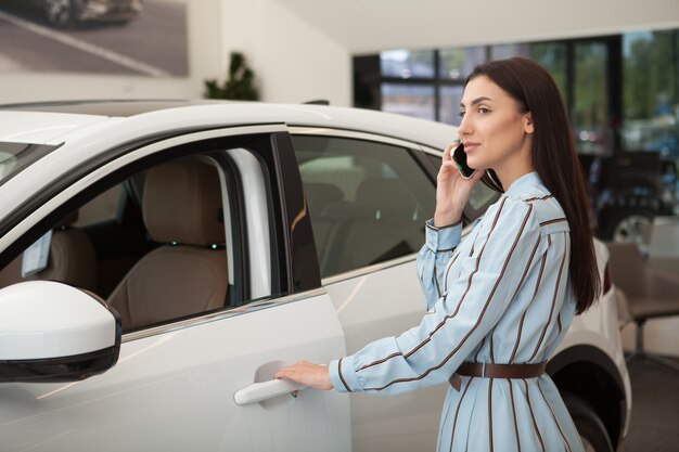 Mooie vrouw die op haar mobiele telefoon praat terwijl ze een auto onderzoekt die te koop is bij de dealer