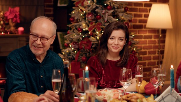 Mooie vrouw die thuis geniet van het kerstdiner met dierbaren terwijl ze hartelijk naar de camera lacht. Feestelijke gelukkige mensen die wintervakantie vieren met traditioneel huisgemaakt eten.