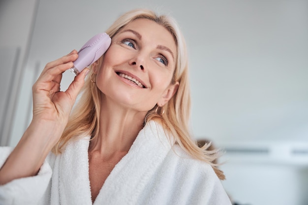 Mooie vrouw die van geweldige tijd geniet terwijl ze een massage op het gezicht van de huid doet met een elektrische gezichtsborstel stimulator