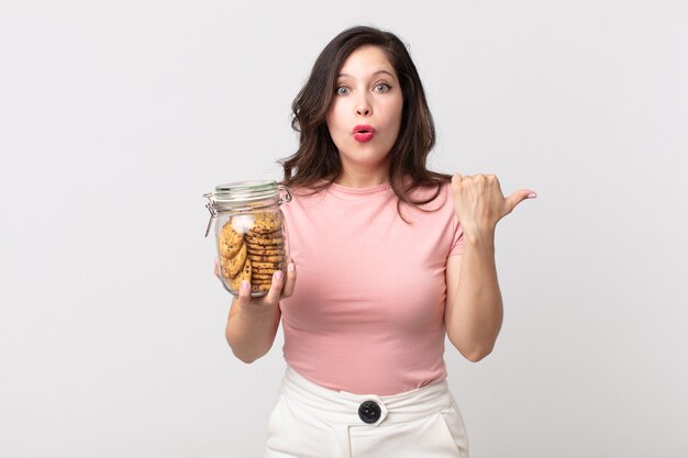 Mooie vrouw die verbaasd kijkt in ongeloof en een glazen fles met koekjes vasthoudt