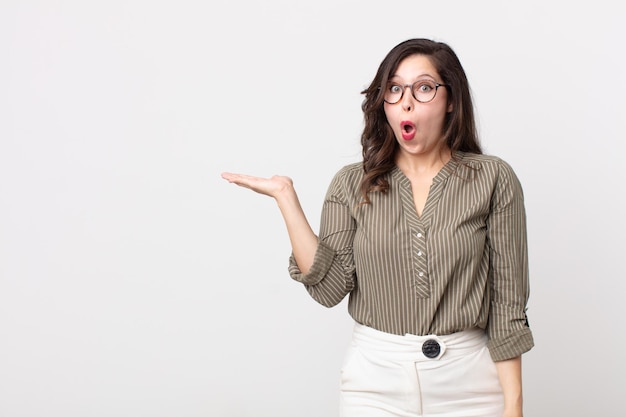 Mooie vrouw die verrast en geschokt kijkt, met open mond terwijl ze een object vasthoudt met een open hand aan de zijkant