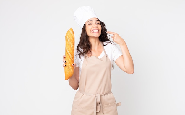 Mooie vrouw die vol vertrouwen glimlacht en naar haar eigen brede glimlach wijst, een schort draagt en een stokbrood vasthoudt