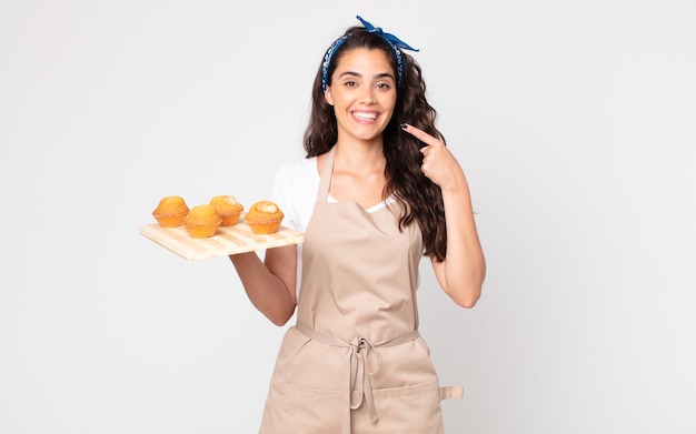 Mooie vrouw die vol vertrouwen glimlacht, wijst naar haar brede glimlach en een dienblad met muffins vasthoudt