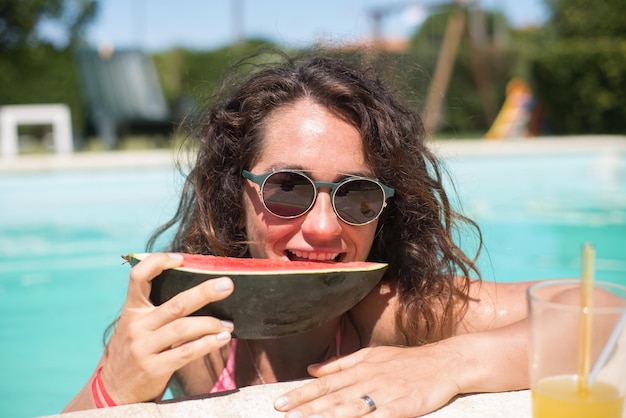 Mooie vrouw die watermeloen in pool eet. Vrouw met donker haar met helder fruit, kijkend naar de camera. Vrije tijd, vriendschap, feestconcept