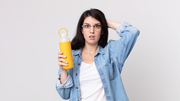 Mooie vrouw die zich gestrest, angstig of bang voelt, met de handen op het hoofd en een koffiethermoskan vasthoudt