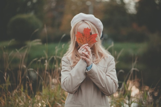 Mooie vrouw die zich verbergt achter rood herfst esdoornblad op de herfst achtergrond
