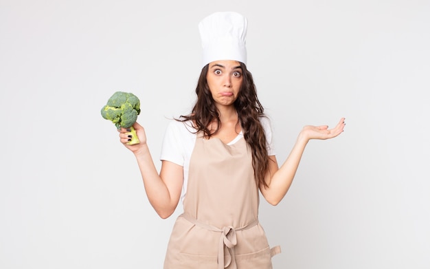 Mooie vrouw die zich verward en verward voelt en twijfelt met het dragen van een schort en het vasthouden van een broccoli