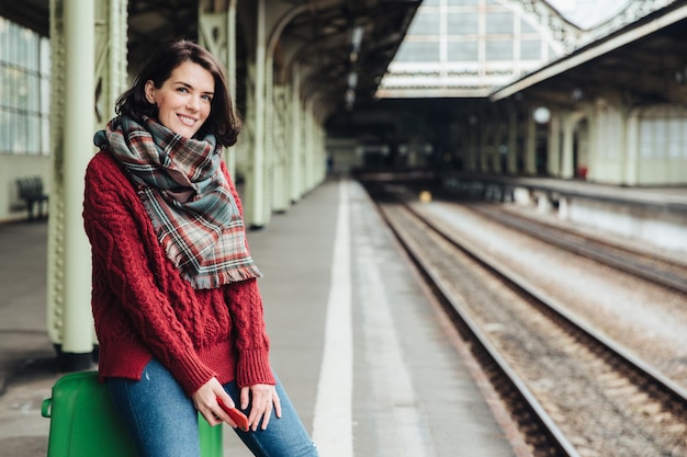 Foto mooie vrouw draagt gebreide trui en sjaal zit op koffer in de buurt van platform houdt smartphone in handen wacht op trein die in een ander land gaat reizen vrouwelijke reiziger met gelukkige uitdrukking