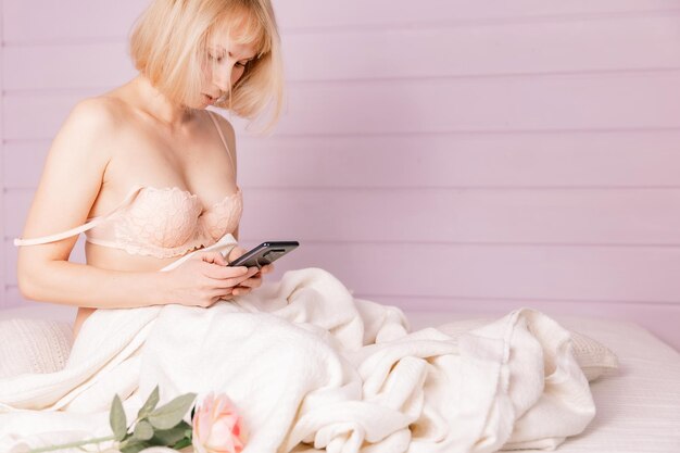 Mooie vrouw dragen luxe lingerie zittend in bed op wit linnen in gezellige flat en het gebruik van smartphone Sexy vrouw zit met telefoon op bed in slaapkamer soft focus Sea...