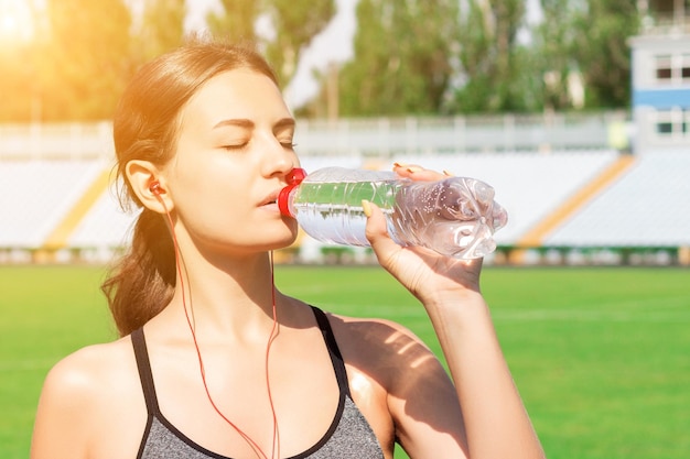 Mooie vrouw drinkt water en luistert naar de muziek op een koptelefoon in het stadion Meisje heeft een pauze na de training