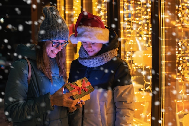 Mooie vrouw en haar zoon in een kerstmuts die een geschenkdoos opent op een verlichte etalageachtergrond