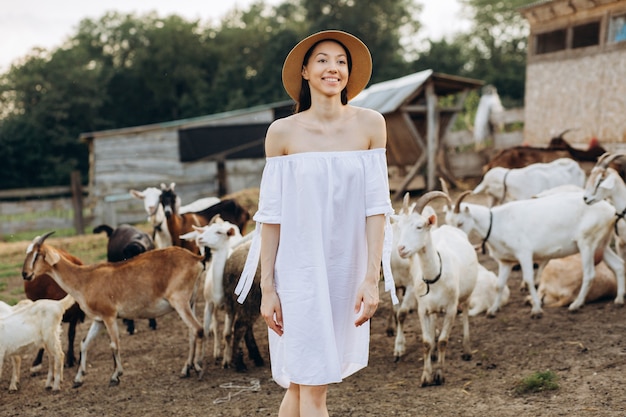 Mooie vrouw en witte jurk en in een beige hoed onder geiten op een eco-boerderij