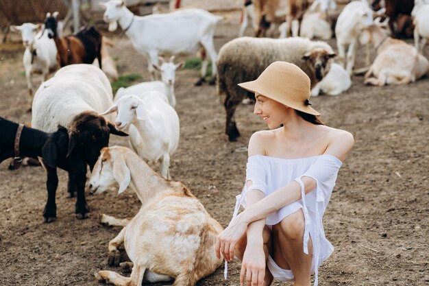 Mooie vrouw en witte jurk en in een beige hoed onder geiten op een eco-boerderij