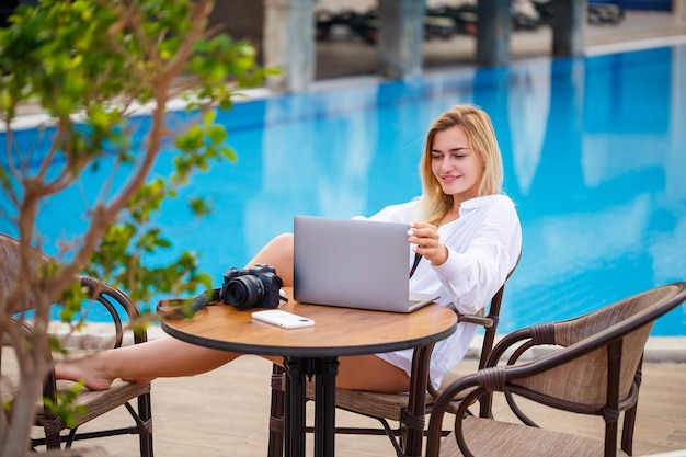 Mooie vrouw fotograaf blogger op vakantie, werken met laptop bij het zwembad. Freelance werken op afstand