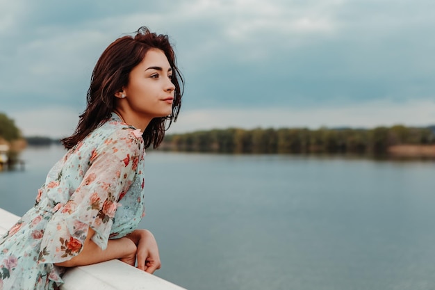 Mooie vrouw gekleed bloemrijke jurk staande op de pier in de buurt van riviermeer humeurig bewolkt weer