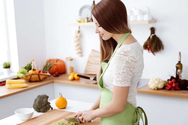 Mooie vrouw gekleed in schort kookt maaltijd in de keuken. Huisvrouw die verse veganistische salade snijdt