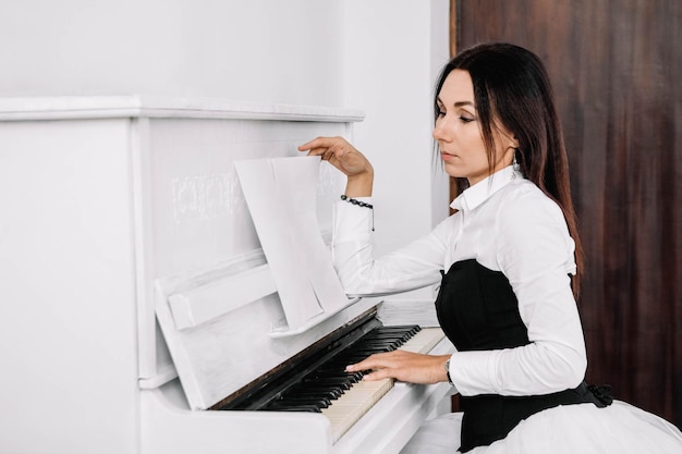 Mooie vrouw gekleed in wit overhemd kijkt naar de muzieknoten en speelt op witte piano