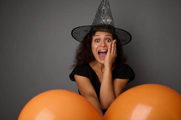 Mooie vrouw gekleed in zwart en tovenaarshoed, kijkt naar de camera, houdt kleurrijke oranje luchtballonnen vast en drukt verbijstering en verrassing uit. Halloween-concept op grijze achtergrond met exemplaarruimte