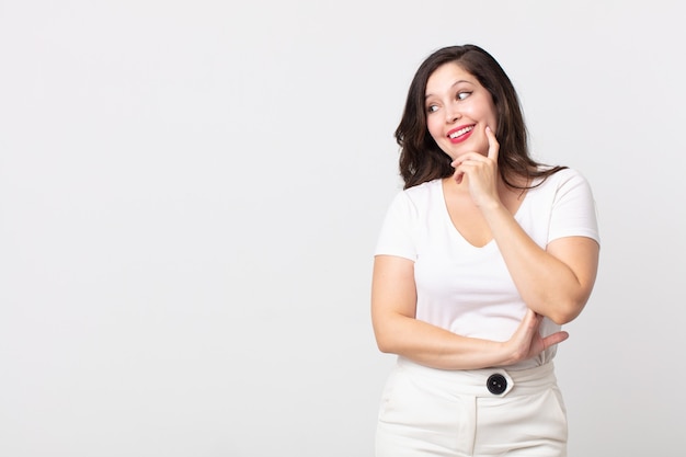 Mooie vrouw glimlachend met een gelukkige, zelfverzekerde uitdrukking met de hand op de kin, zich afvragend en opzij kijkend