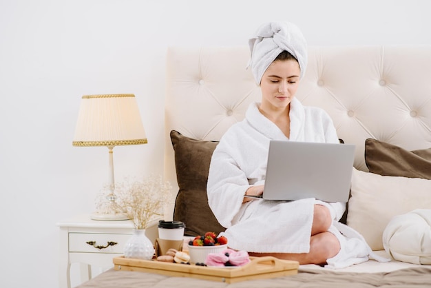 Mooie vrouw in badjas en handdoek op het hoofd met behulp van een laptop op het bed thuis