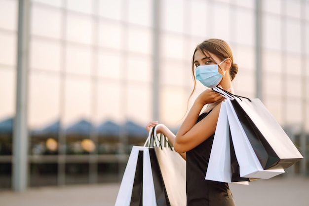 Mooie vrouw in beschermend steriel medisch masker met boodschappentassen in de buurt van winkelcentrum.