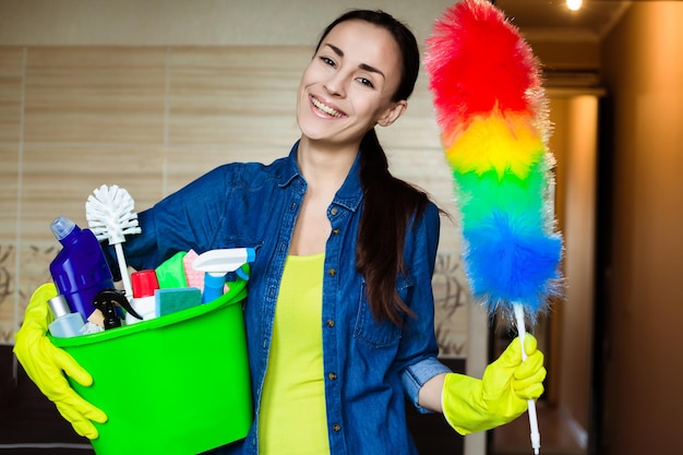 Mooie vrouw in beschermende handschoenen houdt een emmer met dingen om schoon te maken, kijkt naar de camera en glimlacht voor het begin van het schoonmaken van het huis