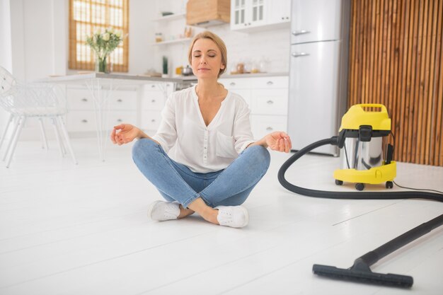 Mooie vrouw in blouse en spijkerbroek in lotushouding op witte vloer thuis na het schoonmaken