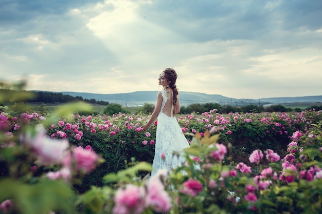 Mooie vrouw in een bloemenpark, tuinrozen. Make-up, haar, een krans van rozen. Lange trouwjurk. Vrouw in boho-stijl