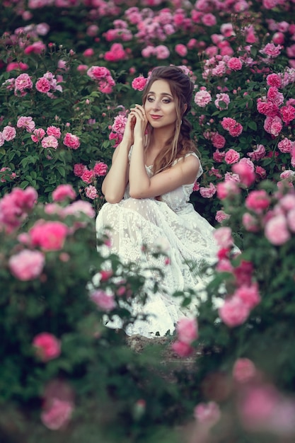 Mooie vrouw in een bloemenpark, tuinrozen. Make-up, haar, een krans van rozen. Lange trouwjurk. Vrouw in boho-stijl