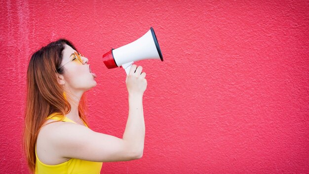 Mooie vrouw in een gele jurk schreeuwt in een megafoon op een rode achtergrond Stijlvolle roodharige dame met zonnebril uitte hun eisen via luidsprekers