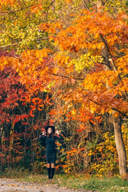 Mooie vrouw in een groene jurk die hoed met haar handen vasthoudt en in de herfst omhoog kijkt naar het steegje in het park