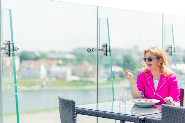 Mooie vrouw in een helder magenta jasje zit op een zomerterras en eet een verse groentesalade. Horizontale foto