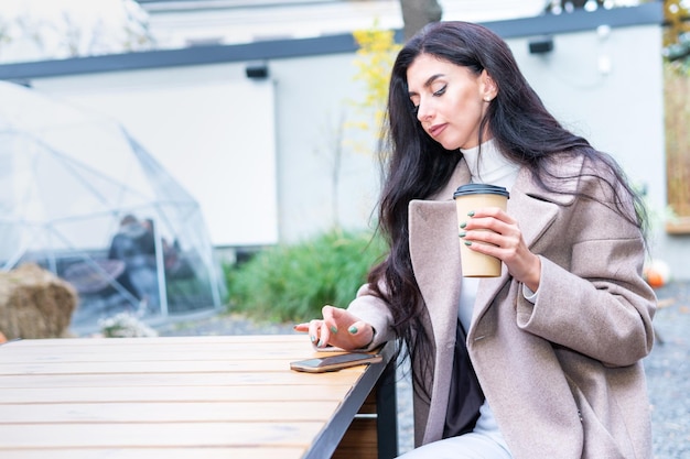 Mooie vrouw in een jas zit aan een tafel in een café, drinkt koffie en gebruikt de telefoon