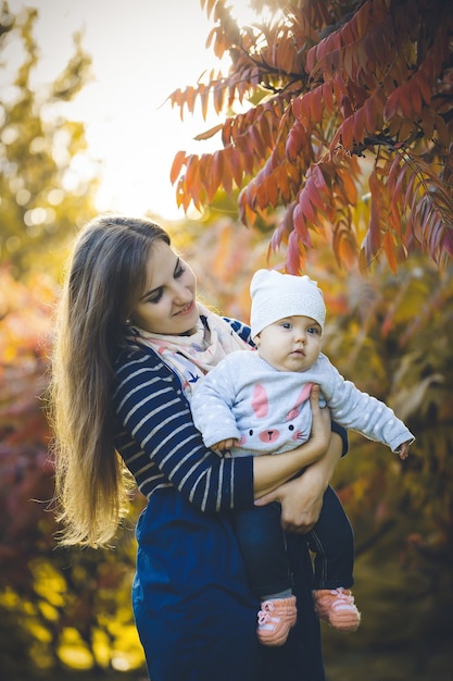 Mooie vrouw in een jurk speelt met een kind in haar armen, staande in de herfst natuur met gevallen bladeren. Het jonge langharige meisje ontspant in het park met rode gele bladeren in de herfst. Gelukkig gezin.